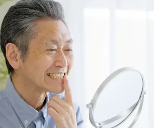 overdentures-1 Man examining teeth in mirror, smiling slightly.