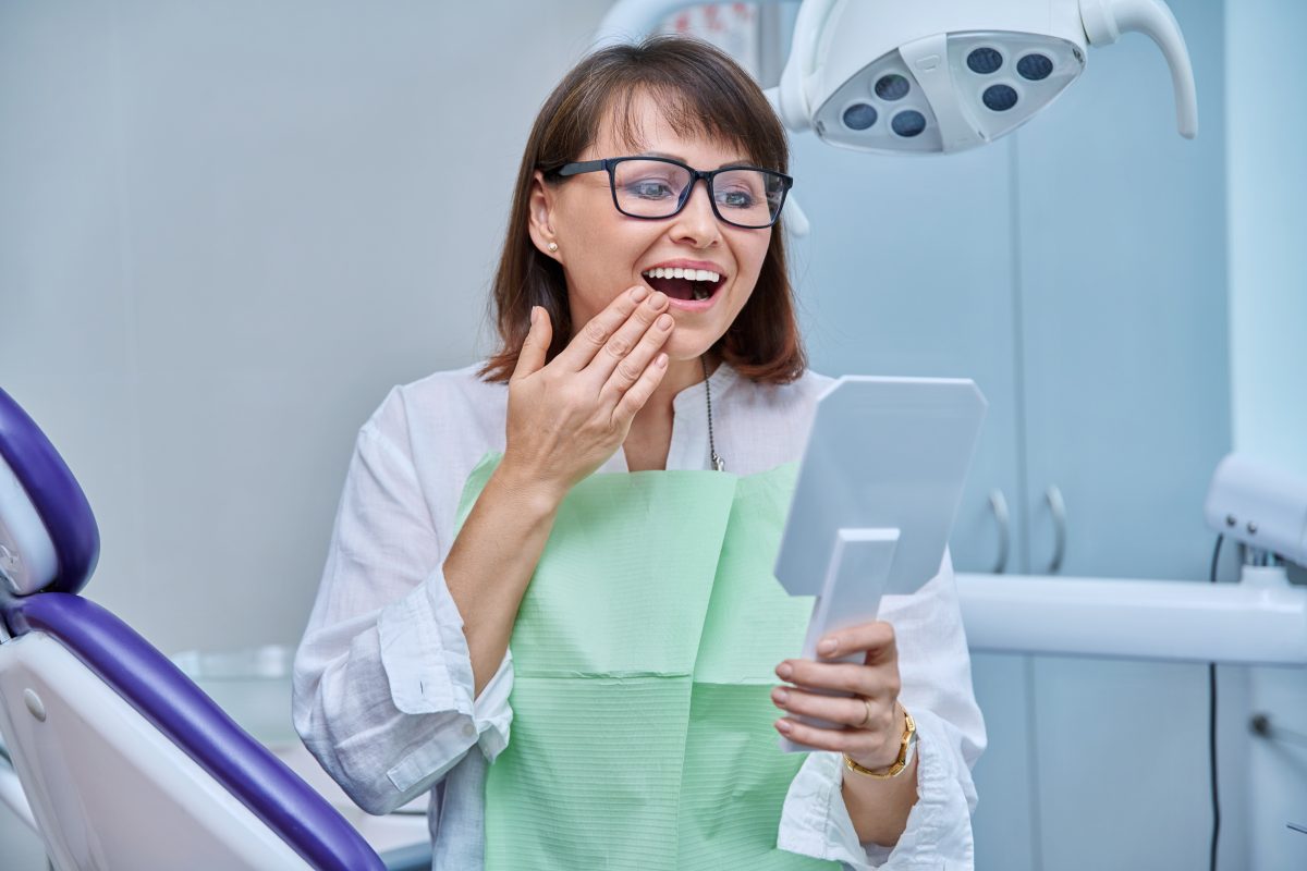 Woman smiling with hand on mouth, holding mirror.